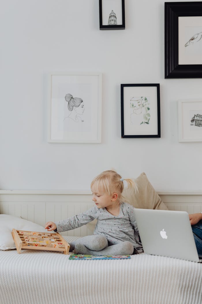 A cute child in a gray sweater sits on a bed playing near a laptop in a cozy room.