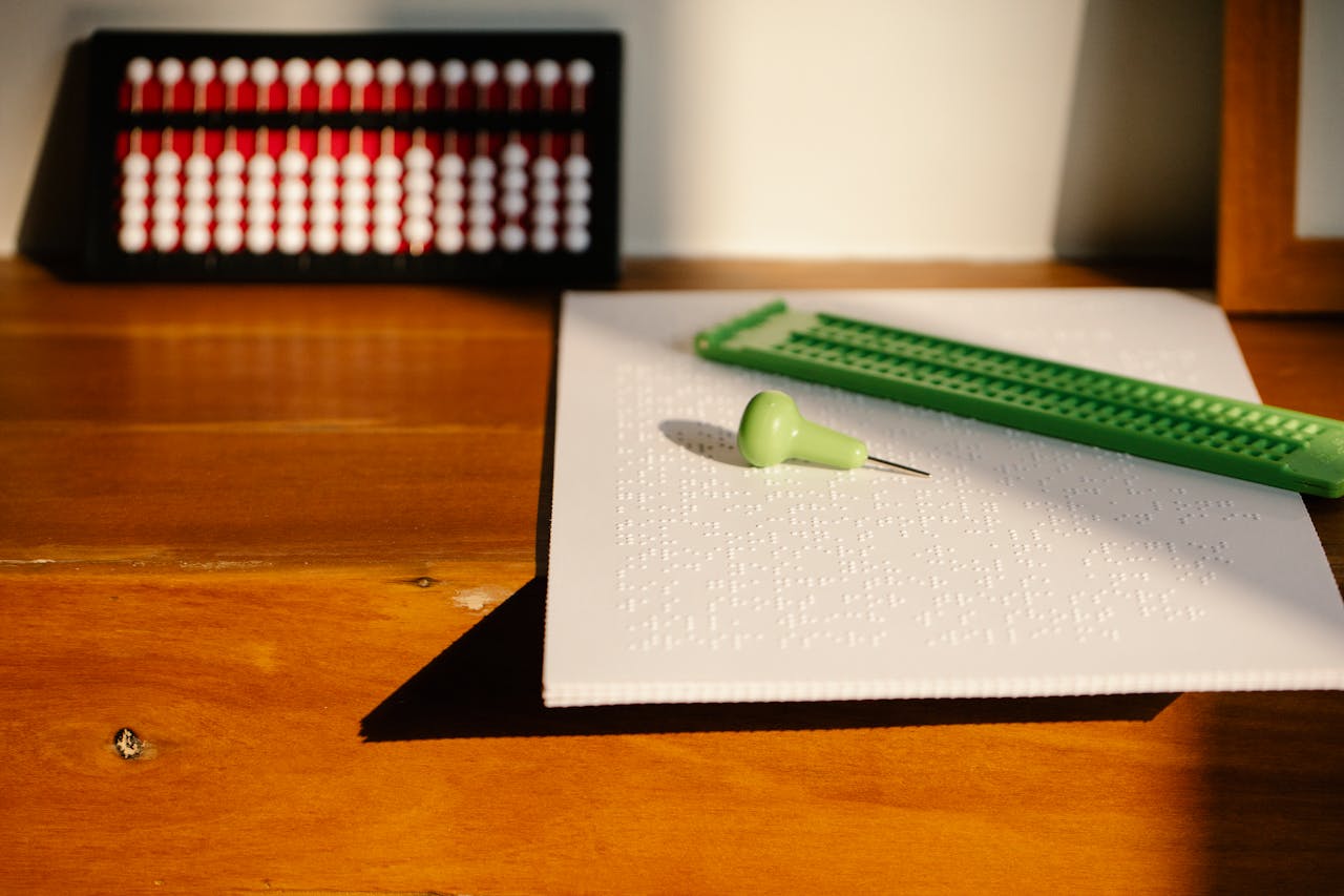Close-up of Braille paper with stylus and abacus on a sunlit wooden desk, highlighting tactile learning tools.
